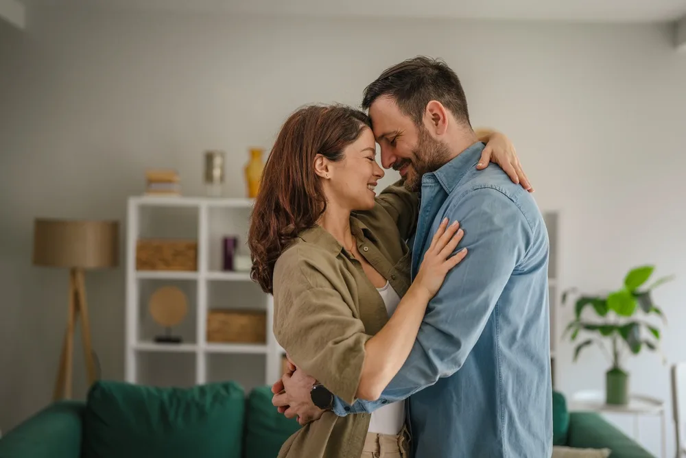Usmievajúci sa partnerský pár sa objíma v obývačke a dotýka sa čelami.
