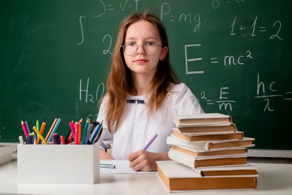 Študentka s okuliarmi sedí pri stole s knihami pred školskou tabuľou s matematickými a fyzikálnymi vzorcami.